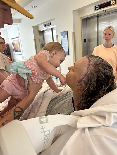 A woman in a hospital bed with scaring on her head holds a young child while smiling with family surrounding her.